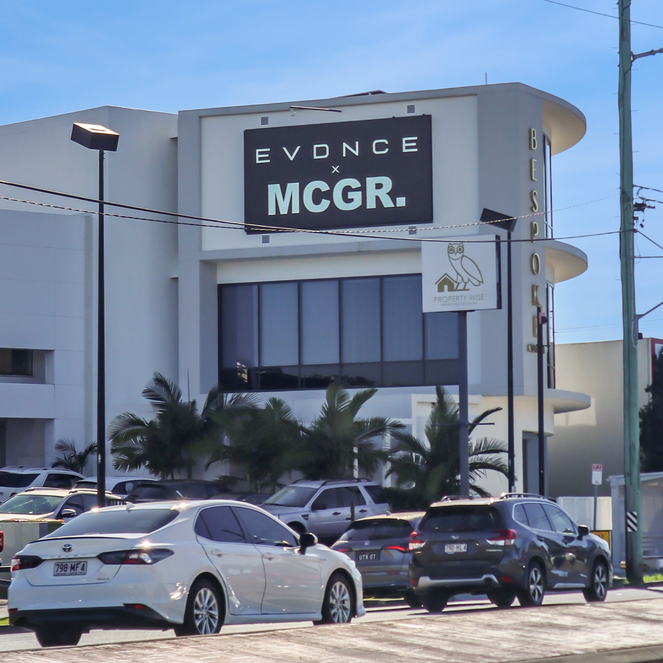 A street view of the Evdance MCGR. building under a bright blue sky, with several cars parked in front and a road in the foreground.
