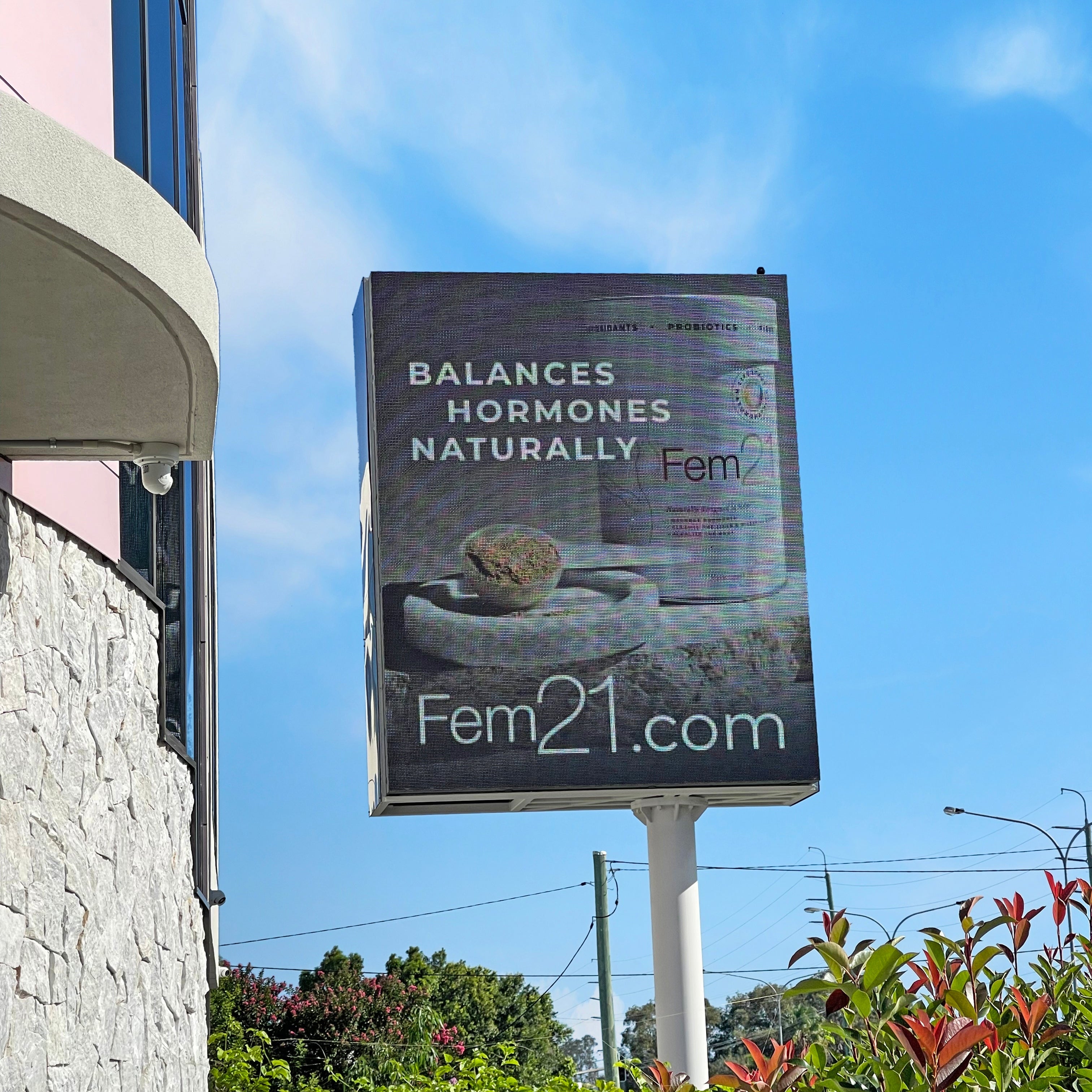 Billboard advertising 'Fem21' product with text 'Balances Hormones Naturally' against a blue sky.