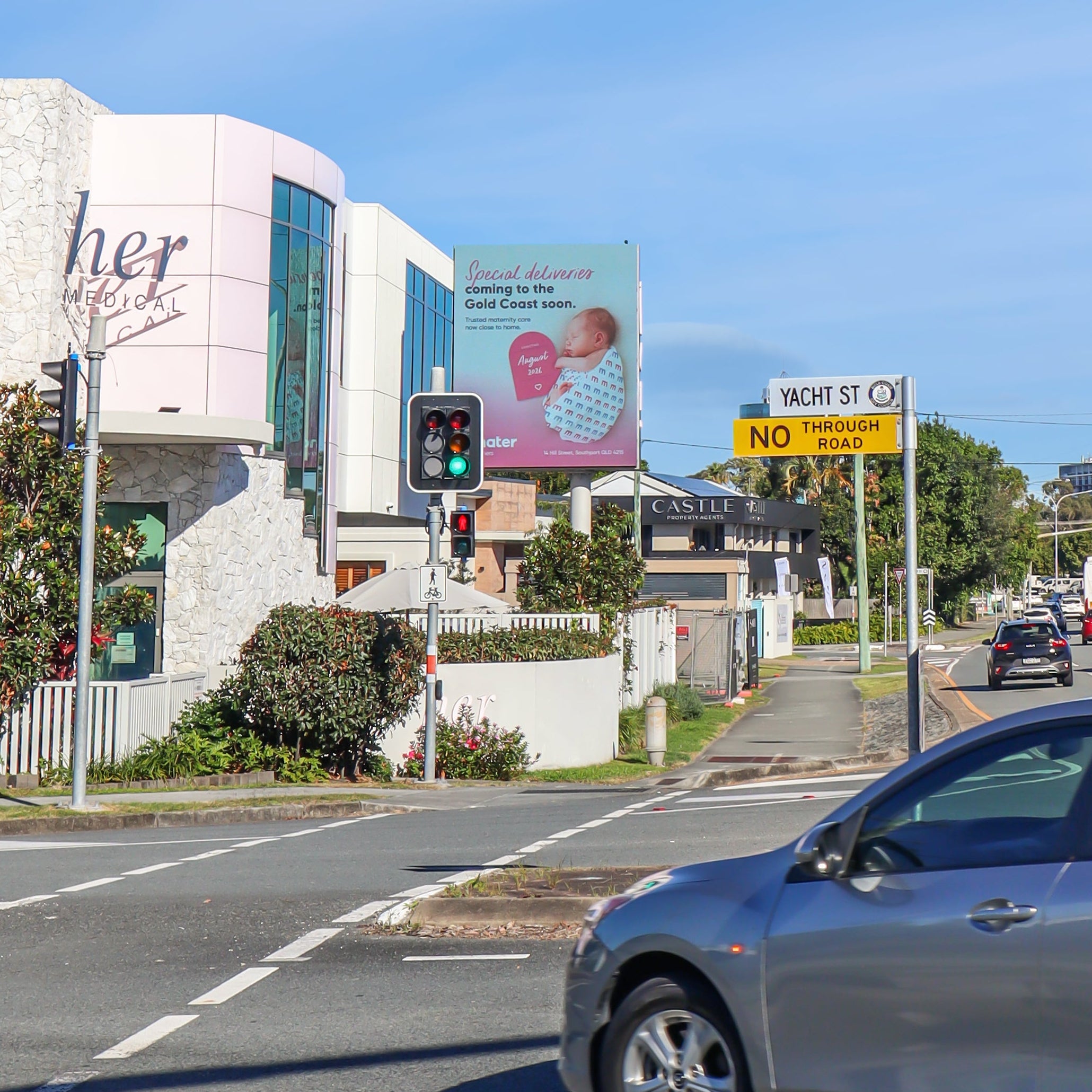 A silver hatchback car driving on a city road under a bright blue sky with light clouds, with buildings, billboards, and other vehicles in the background.