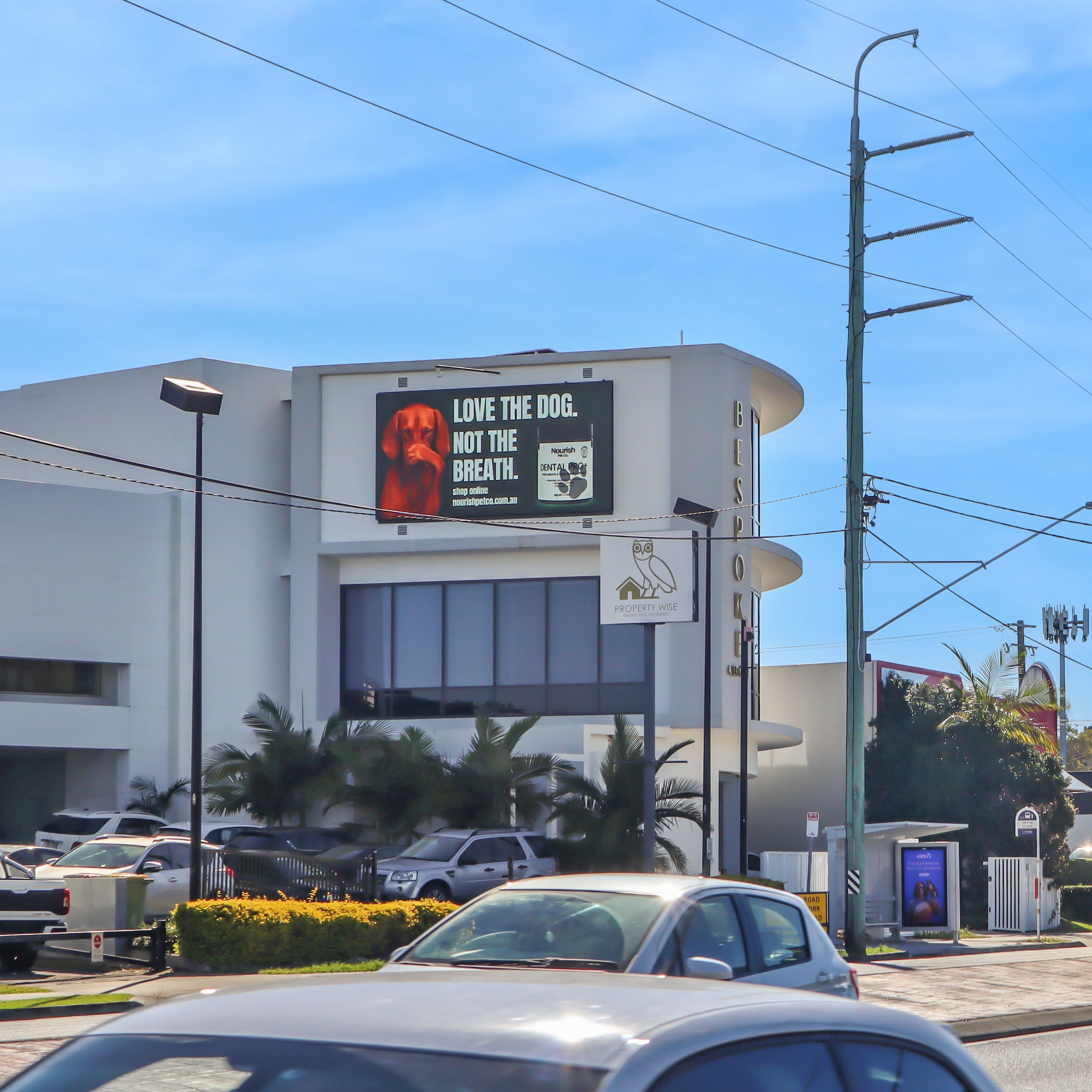 A sunny street scene with multiple cars on a road, utility poles, overhead wires, and a white commercial building featuring a large green sign with a dog covering it's nose that reads 'LOVE THE DOG. NOT THE BREATH'